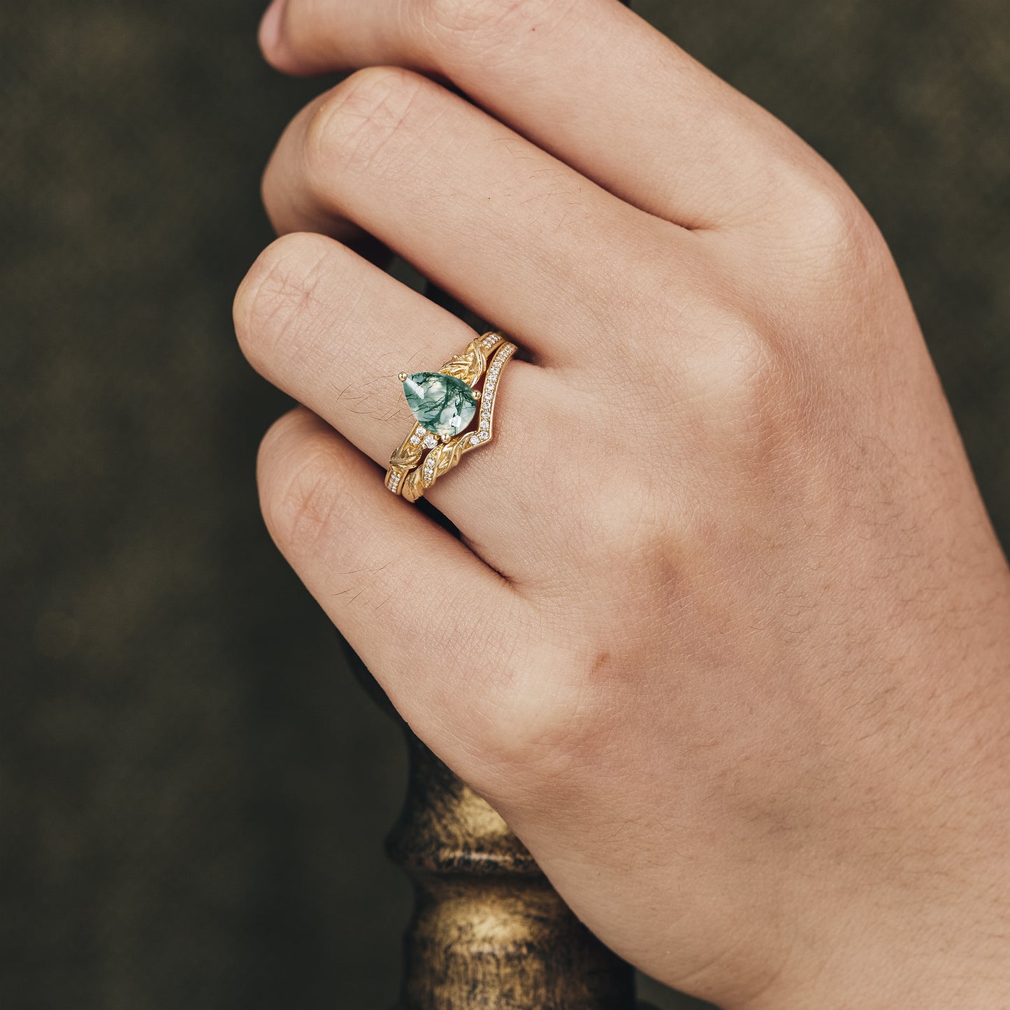 Hand displaying asymmetric pear-shaped moss agate engagement ring with gold design.